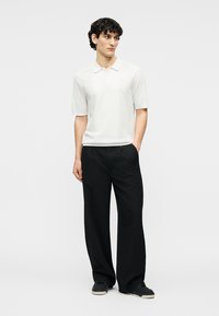 Άνδρας όρθιος, φοράει λευκό πόλο μπλουζάκι με κοντά μανίκια, μαύρο παντελόνι με φαρδύ πόδι και μαύρα αθλητικά παπούτσια, μπροστά σε απλό φωτεινό φόντο.