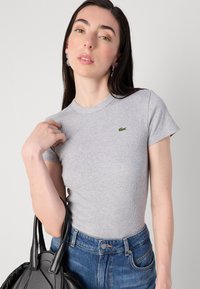 Modelis su ilgais plaukais vilki pilką prigludusią marškinėlių su mažu žaliu Lacoste logotipu ir džinsais, laiko juodą rankinę. Neutralus fonas.