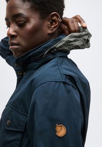 Personne à la peau foncée ajustant la capuche d'une veste bleu foncé avec un écusson de logo renard sur la manche, portant trois boucles d'oreilles à l'oreille gauche, devant un fond uni.