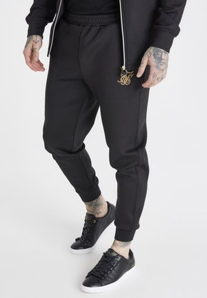 Schwarze Jogginghosen mit elastischem Bund, seitlichen Taschen und goldfarbenem Logoakzent. Kombiniert mit schwarzen strukturierten Sneakers.