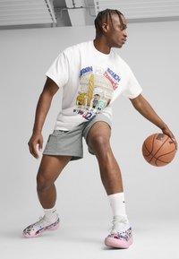 Hvid grafisk t-shirt med farverigt tegneserie-design, grå shorts og multikolore sneakers, mens han holder en basketball i en dynamisk positur.