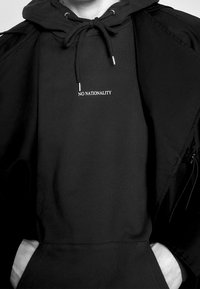 Czarna bluza z kapturem i sznurkami, z białym haftem z napisem "NO NATIONALITY". Połączona z czarną kurtką wierzchnią.