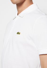 Polo blanc en tissu texturé, avec un col classique, trois boutons, et un petit logo crocodile vert et or sur la poitrine.