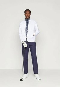 Balts bomber jakas, rakstains tumši zilas krāsas krekls, tumši zilas bikses, baltas golfa cimdi, tur rokās golfa nūju. Apavi ir balti sporta apavi.