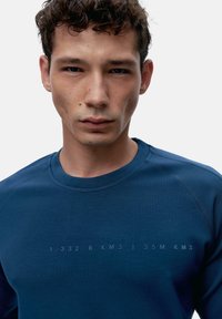 Modra bluza s krojem iz okroglega vratu, izdelana iz gladkega materiala; ima tiskano besedilo na prsih, prilegajoč dizajn in raglan rokave.