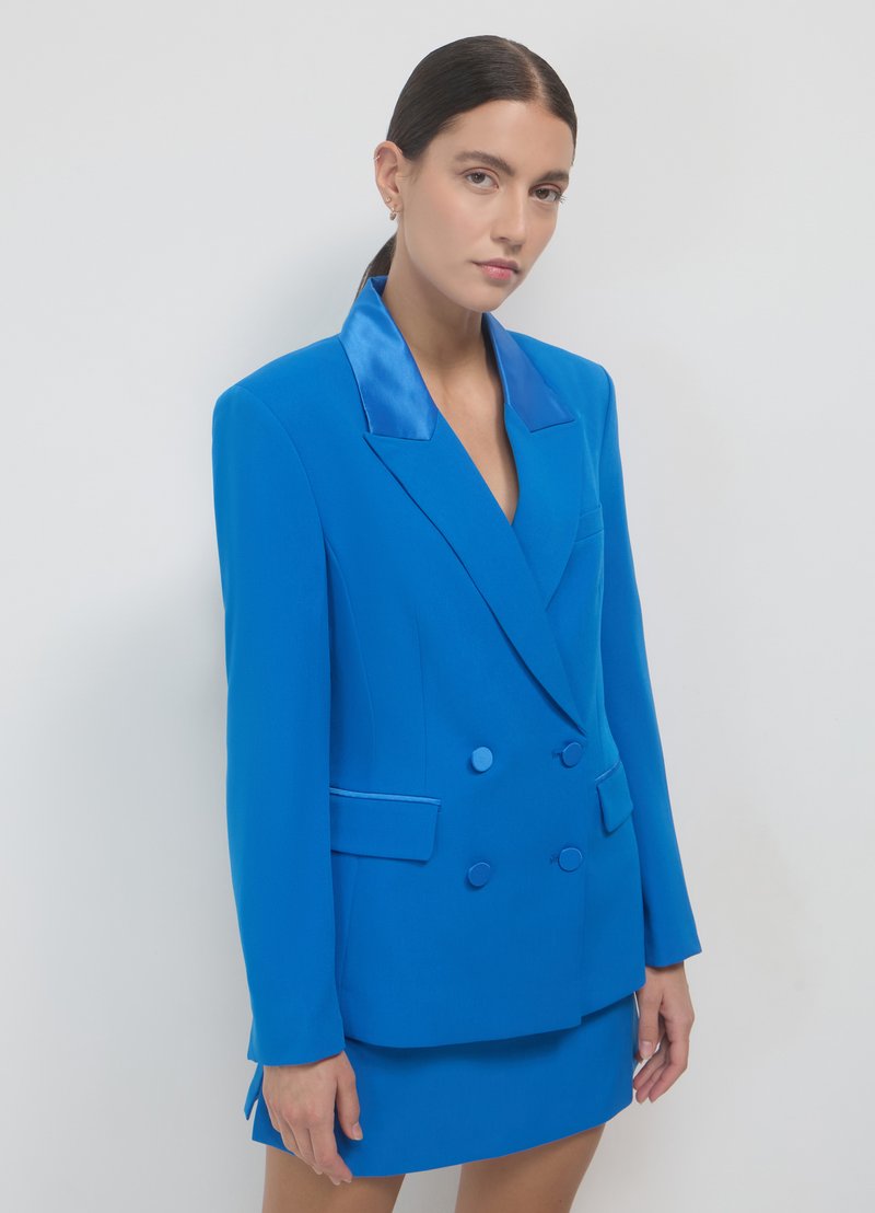 Blazer doppiopetto color blu brillante con dettagli in satin, rever a incavo e tasche con patta. Abbinato a una minigonna coordinata. Tessuto liscio.