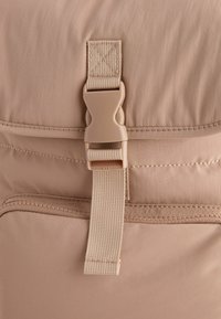 Beige szövet hátizsák rész nylon csattal és pánttal. Tartalmaz vízszintes varrási részleteket és sima külső textúrát.