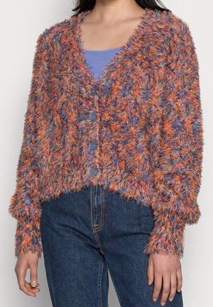Person trägt eine mehrfarbige, flauschige Strickjacke mit blauen Knöpfen über einem lila Oberteil und dunkelblauen Jeans.