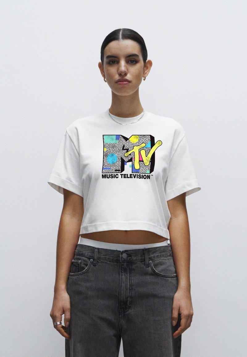 White cropped t-shirt featuring a graphic design with black, yellow, and colorful elements, accompanied by "MUSIC TELEVISION" text below.