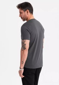 Šedé bavlněné tričko s krátkým rukávem, kulatým výstřihem a slim fit střihem. Viditelné tetování na ruce, nošeno s černými kalhotami.