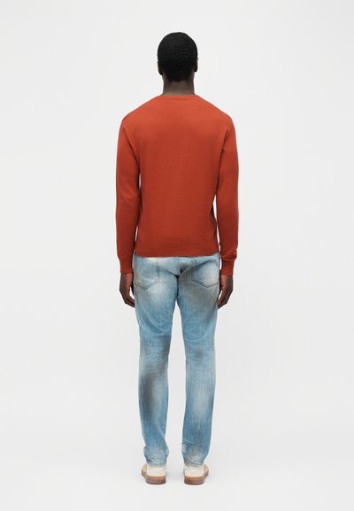 Homme portant un pull à manches longues couleur rouille, un jean bleu clair délavé et des chaussures blanches, debout de dos sur un fond blanc uni.