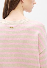 Μπεζ πλεκτό πουλόβερ με οριζόντιες ροζ ρίγες, ριμπ λαιμόκοψη και χρυσή ετικέτα στο πίσω μέρος. Απαλή υφή και άνετη εφαρμογή.