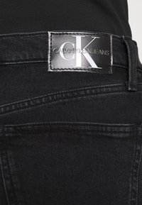 Musta denimfarkut, joissa on hopeinen "CALVIN KLEIN JEANS" -logo nahkalapussa. Mallissa on näkyvät saumat ja klassinen leikkaus.