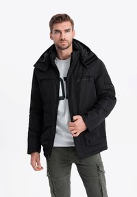 Černá izolovaná bunda s vysokým límcem a odnímatelnou kapucí. Obsahuje zipovou uzávěrku a více kapes. Nositá přes šedou mikinu.