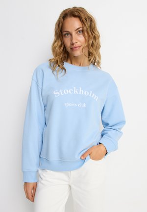 Frau mit welligem blondem Haar, die einen hellblauen Pullover mit dem Schriftzug "Stockholm Sports Club" und weiße Hosen trägt, posiert vor weißem Hintergrund.