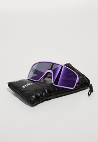 Lunettes de soleil oversized violettes avec une finition mate et des verres dégradés, posées sur un étui rembourré noir avec un logo.