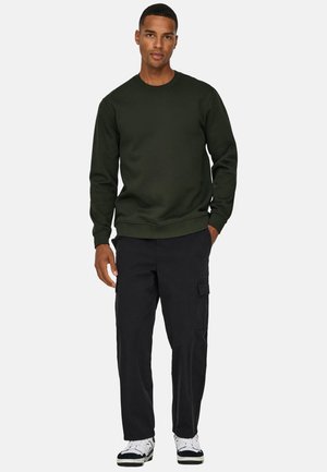 Mies seisoo kädet taskuissa yllään tummanvihreä collegepaita, mustat cargohousut ja valkoiset tennarit, vaaleanharmaata taustaa vasten.