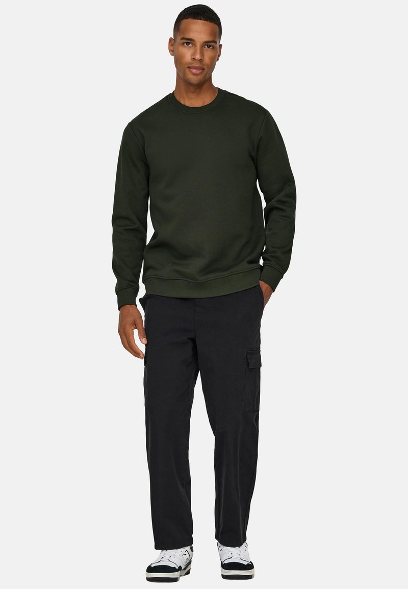 Mann, der einen dunkelgrünen Pullover, schwarze Cargohosen und weiße Turnschuhe trägt, mit den Händen in den Taschen vor einem hellgrauen Hintergrund steht.