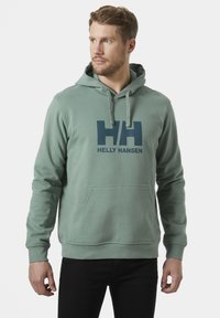 Sudadera verde hecha de tela suave, con un bolsillo tipo canguro y cordones ajustables. Gran logo azul de Helly Hansen impreso en la parte frontal.