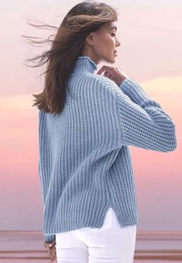 Gaiši zila ribota adīta džemperis ar augstu apkakli, sānu caurumiem un garām piedurknēm, pieskaņots baltām pieguļošām biksēm. Redzama mīksta tekstūra.