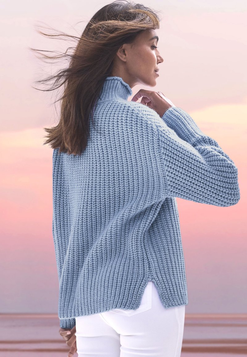 Gaiši zila ribota adīta džemperis ar augstu apkakli, sānu caurumiem un garām piedurknēm, pieskaņots baltām pieguļošām biksēm. Redzama mīksta tekstūra.