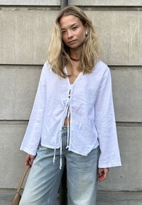Jeune femme aux longs cheveux blonds portant un haut blanc noué devant, un jean bleu clair, un collier de perles rouges, tenant un sac à main beige, debout à l'extérieur.