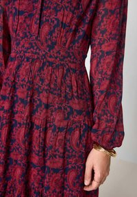 Abito rosso e blu con motivo floreale intricato, tessuto plissettato, maniche lunghe e vita aderente. Bracciali dorati al polso.