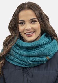 Bufanda infinity de punto en color verde azulado con un patrón texturizado. Tejido suave y cálido con una etiqueta de cuero. Llevada sobre una chaqueta oscura.