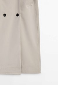 Ourlet de trench-coat beige avec deux boutons noirs et des détails cousus le long du bord inférieur.