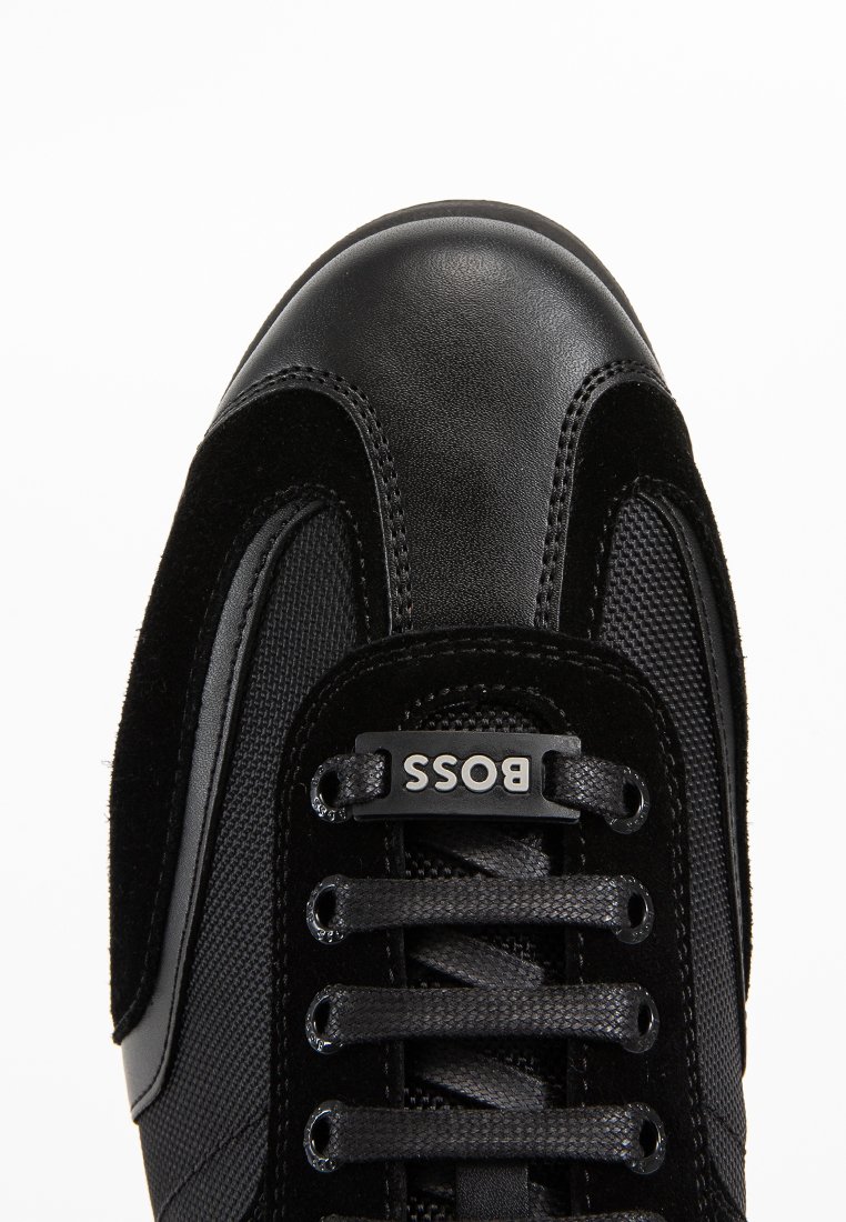 Baskets noires avec un bout en cuir lisse, des côtés en maille noire texturée et des accents en daim. Elles possèdent des lacets plats noirs et une étiquette logo.