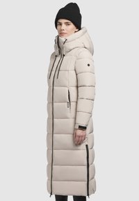 Long, beige puffer coat with a hood, featuring black zipper closures and adjustable drawstrings. Textured, quilted pattern throughout.