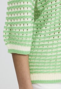 Detailný záber na rukáv zeleno-bieleho pásikavého pleteného svetra s textúrovanými horizontálnymi líniami na ramene osoby.