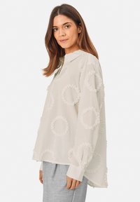 White blouse with a collar and button placket. Features textured, circular embroidery throughout. Long sleeves with cuff detailing.