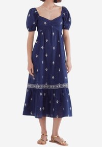 Robe longue bleu marine à manches bouffantes en tissu léger, ornée de motifs floraux blancs et d'une bordure en dentelle à l'ourlet. Sandales plates portées.