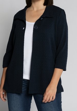 Marineblå strukturerad cardigan med trekvartsärmar, som har en enda svart knappstängning och öppen framsidesdesign, lagd över en vit skjorta.