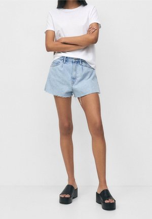 Shorts en denim bleu clair, avec un ourlet effiloché, taille haute, accompagnés d'un t-shirt blanc uni et de sandales à plateforme noires.