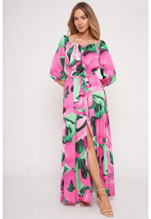 Frau in einem langen Kleid mit einem Muster in Pink, Grün und Schwarz, mit Puffärmeln und einem hohen Beinschlitz, trägt hautfarbene Sandalen mit hohen Absätzen.