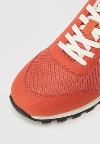 Zapatilla naranja con parte superior de malla, cordones blancos y suela de goma de contraste con detalles texturizados. Punta redondeada y acentos cosidos.