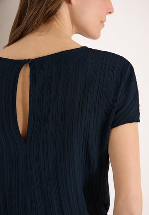 Femme portant un haut noir à manches courtes texturé avec une petite ouverture en forme de goutte et une fermeture par bouton dans le haut du dos.