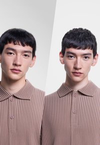 Polo shirt marrone a coste con colletto ripiegato e due bottoni. Il tessuto ha un aspetto texturizzato. Sfondo diviso in grigio chiaro e bianco.