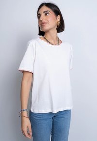 T-shirt bianco in cotone con maniche corte, caratterizzato da cuciture a contrasto rosse lungo il colletto e le maniche. Abbinato a jeans blu.