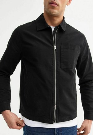 Chaqueta negra con cremallera hecha de algodón, que presenta un cuello tipo solapa, un bolsillo en el pecho y una textura lisa. Mangas largas con dobladillo recto.