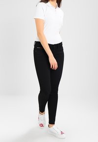 Žena kráčí, má na sobě bílou polo košili, černé skinny džíny a bílé tenisky s červenými květinovými výšivkami.