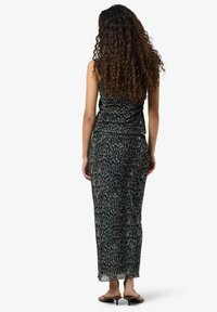 Donna con lunghi capelli ricci, che indossa un top senza maniche con stampa leopardata e una lunga gonna coordinata, in piedi con i piedi in sandali neri con tacco.
