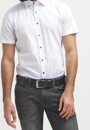 Mand iført en tætsiddende hvid skjorte med korte ærmer og knapper, stoppet ned i mørkegrå jeans med sort bælte.