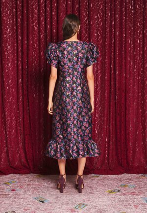 Femme debout face à un rideau floral rouge, portant une robe midi fleurie foncée avec des manches bouffantes et un ourlet à volants, chaussures à talons hauts violettes.