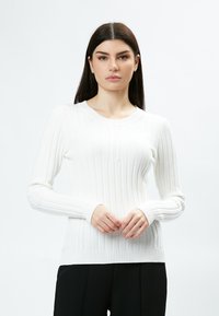 Pullover bianco a coste con maniche lunghe, scollo rotondo, dettagli testurizzati e silhouette aderente, abbinato a pantaloni neri.
