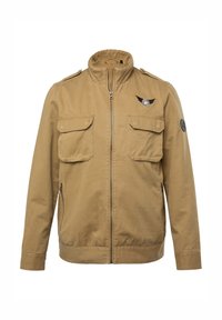 Giacca khaki con colletto alto, zip frontale, due tasche sul petto e un'emblema sulla spalla sinistra. Materiale in cotone con una texture liscia.