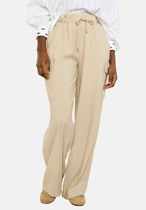 Beige weite Hosen mit Tunnelzug an der Taille und seitlichen Cargotaschen, kombiniert mit braunen Mokassins und einer weißen Langarmbluse mit ausgeschnittenen Manschetten.