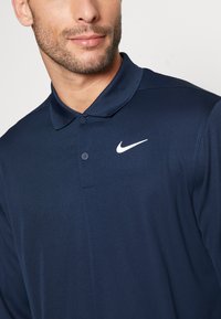 Polo shirt da Nike em azul-marinho, de mangas curtas, com uma placket de dois botões, feito de um tecido suave, apresentando o logotipo swoosh branco no lado esquerdo do peito.